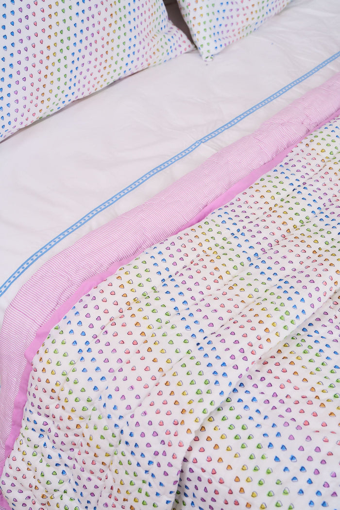 A neatly made twin bed features the Roller Rabbit Disco Hearts Quilt with white bedding and a colorful small heart pattern, complemented by a folded lavender blanket, pink lamp, and books on a white nightstand.
