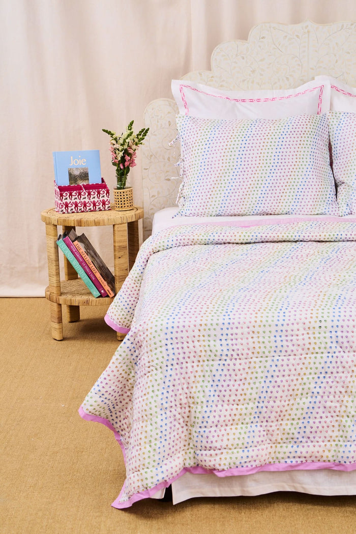 A neatly made twin bed features the Roller Rabbit Disco Hearts Quilt with white bedding and a colorful small heart pattern, complemented by a folded lavender blanket, pink lamp, and books on a white nightstand.