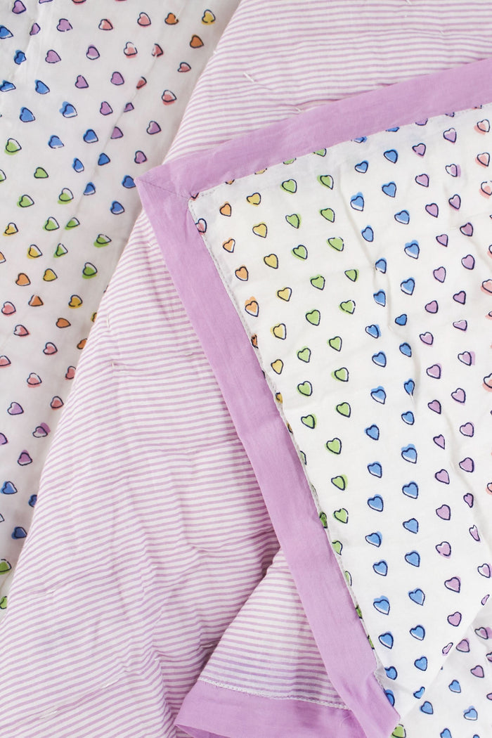 A neatly made twin bed features the Roller Rabbit Disco Hearts Quilt with white bedding and a colorful small heart pattern, complemented by a folded lavender blanket, pink lamp, and books on a white nightstand.