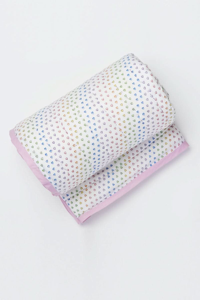 A neatly made twin bed features the Roller Rabbit Disco Hearts Quilt with white bedding and a colorful small heart pattern, complemented by a folded lavender blanket, pink lamp, and books on a white nightstand.