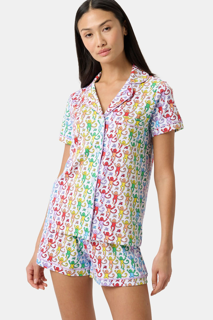 A woman stands barefoot in a short-sleeved Roller Rabbit Disco Monkey Polo Pajama set with matching shorts, featuring a playful, multicolored monkey print on a white background.