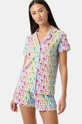 A woman stands barefoot in a short-sleeved Roller Rabbit Disco Monkey Polo Pajama set with matching shorts, featuring a playful, multicolored monkey print on a white background.