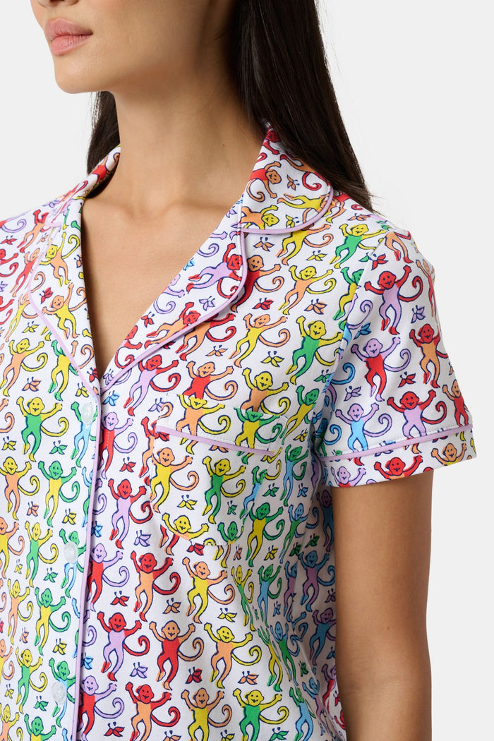 A woman stands barefoot in a short-sleeved Roller Rabbit Disco Monkey Polo Pajama set with matching shorts, featuring a playful, multicolored monkey print on a white background.
