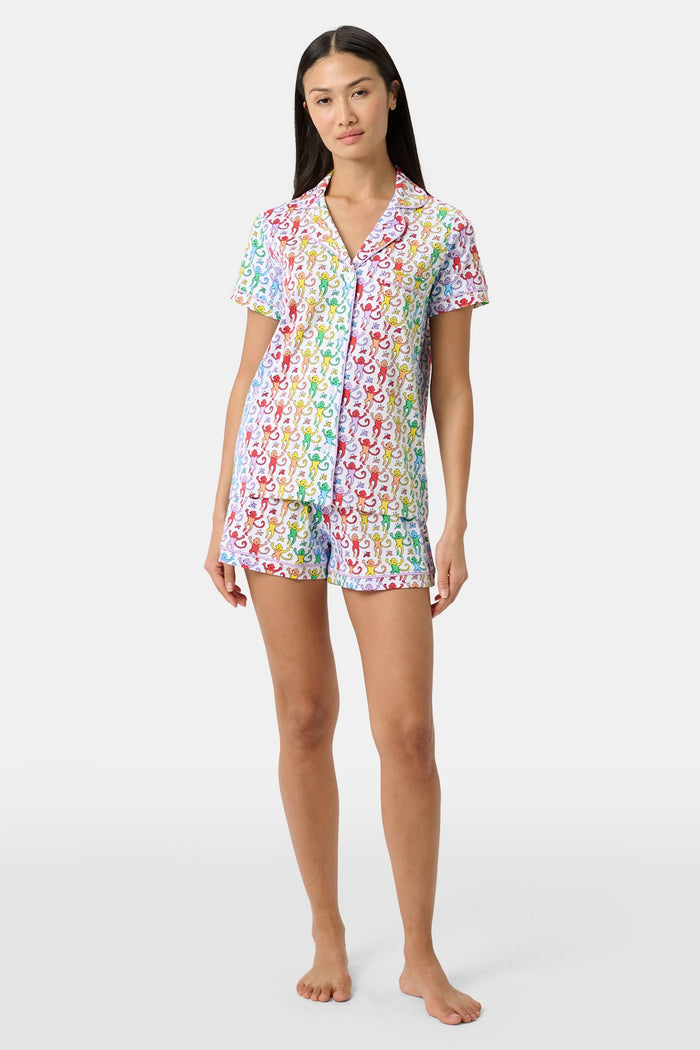 A woman stands barefoot in a short-sleeved Roller Rabbit Disco Monkey Polo Pajama set with matching shorts, featuring a playful, multicolored monkey print on a white background.
