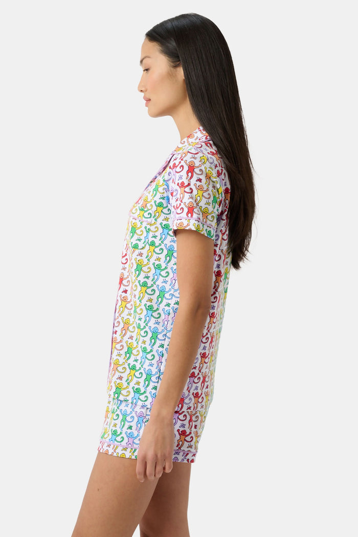 A woman stands barefoot in a short-sleeved Roller Rabbit Disco Monkey Polo Pajama set with matching shorts, featuring a playful, multicolored monkey print on a white background.