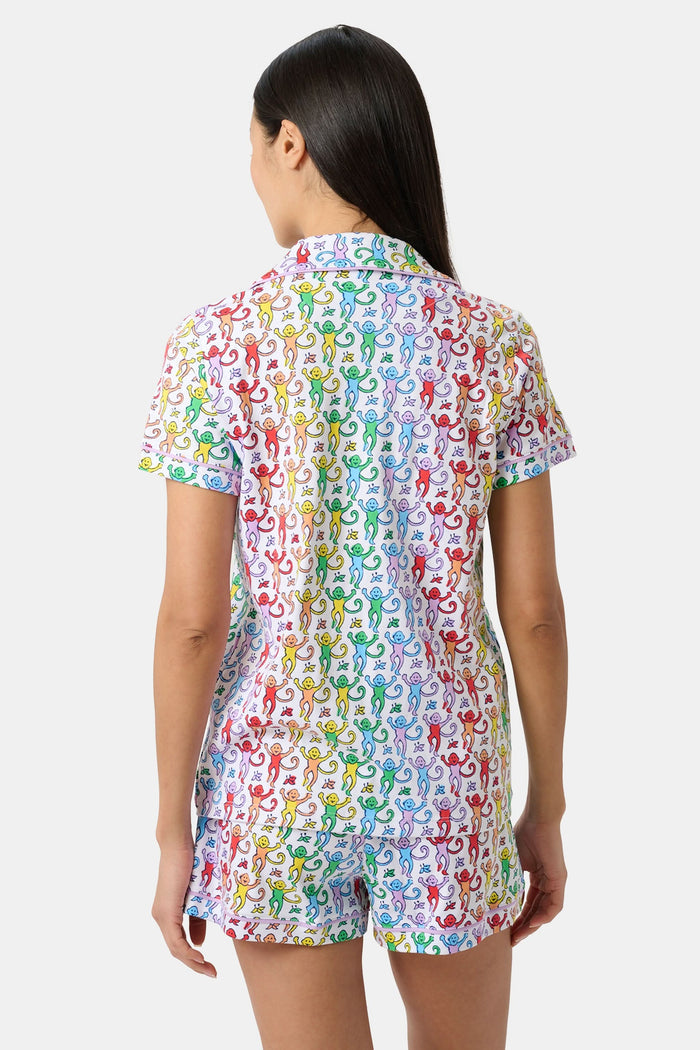A woman stands barefoot in a short-sleeved Roller Rabbit Disco Monkey Polo Pajama set with matching shorts, featuring a playful, multicolored monkey print on a white background.