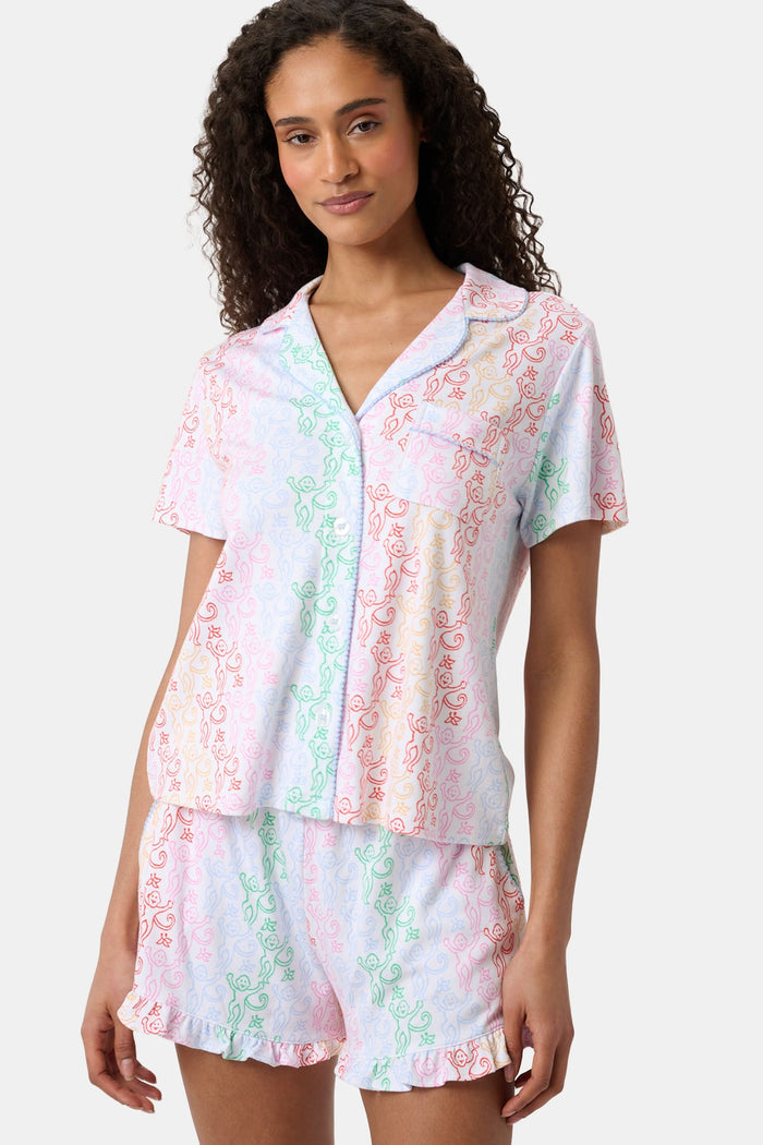 A woman with curly hair, standing and smiling slightly, wears Roller Rabbit’s Disco Monkey Wyatt Polo Pajamas: a short-sleeve button-up top and matching ruffled shorts featuring a pastel multicolor abstract pattern on white.