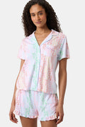 A woman with curly hair, standing and smiling slightly, wears Roller Rabbit’s Disco Monkey Wyatt Polo Pajamas: a short-sleeve button-up top and matching ruffled shorts featuring a pastel multicolor abstract pattern on white.