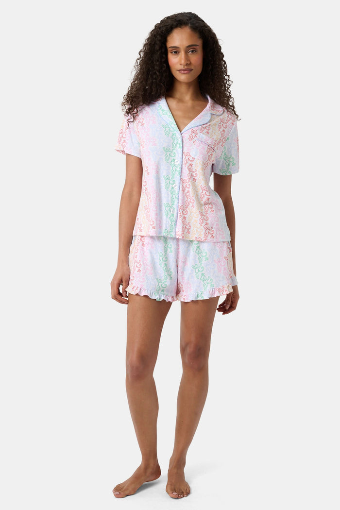 A woman with curly hair, standing and smiling slightly, wears Roller Rabbit’s Disco Monkey Wyatt Polo Pajamas: a short-sleeve button-up top and matching ruffled shorts featuring a pastel multicolor abstract pattern on white.