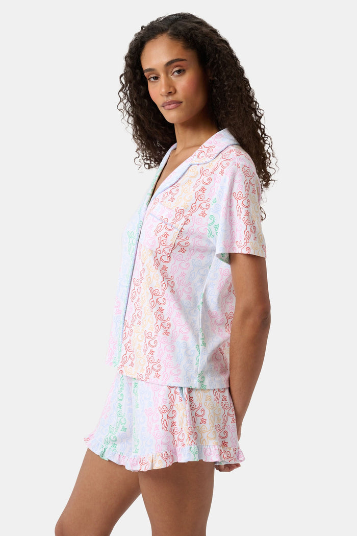 A woman with curly hair, standing and smiling slightly, wears Roller Rabbit’s Disco Monkey Wyatt Polo Pajamas: a short-sleeve button-up top and matching ruffled shorts featuring a pastel multicolor abstract pattern on white.