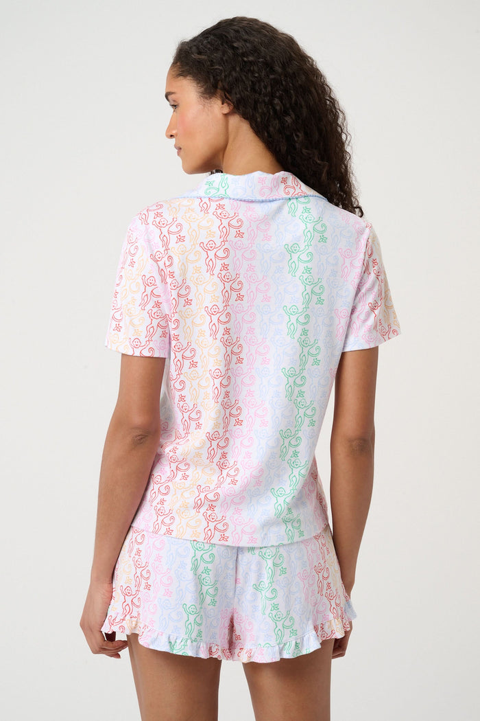 A woman with curly hair, standing and smiling slightly, wears Roller Rabbit’s Disco Monkey Wyatt Polo Pajamas: a short-sleeve button-up top and matching ruffled shorts featuring a pastel multicolor abstract pattern on white.
