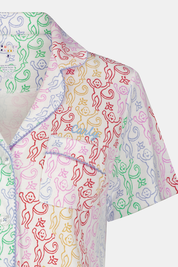 A woman with curly hair, standing and smiling slightly, wears Roller Rabbit’s Disco Monkey Wyatt Polo Pajamas: a short-sleeve button-up top and matching ruffled shorts featuring a pastel multicolor abstract pattern on white.