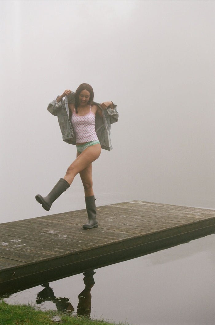 A woman stands against a light background, hands behind her back, wearing Roller Rabbit’s Ditsy Daisy Pointelle Carmine Cami and matching underwear with a small red heart pattern and lace trim.