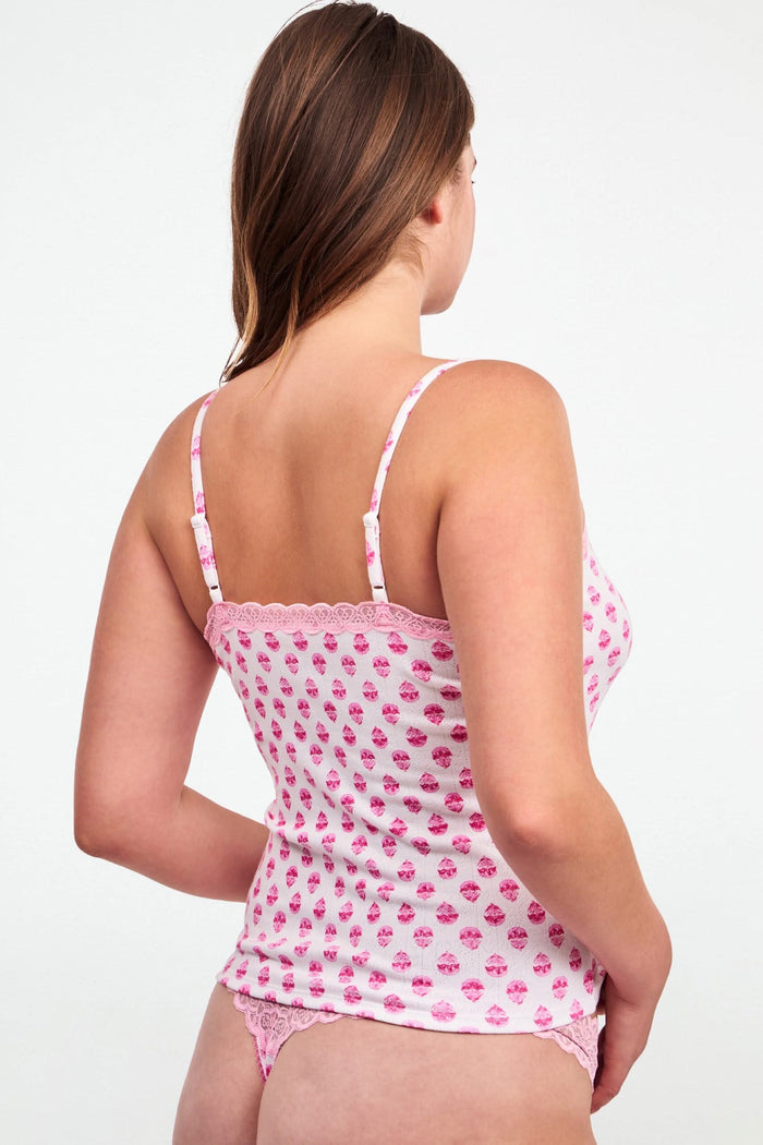 A woman stands against a light background, hands behind her back, wearing Roller Rabbit’s Ditsy Daisy Pointelle Carmine Cami and matching underwear with a small red heart pattern and lace trim.
