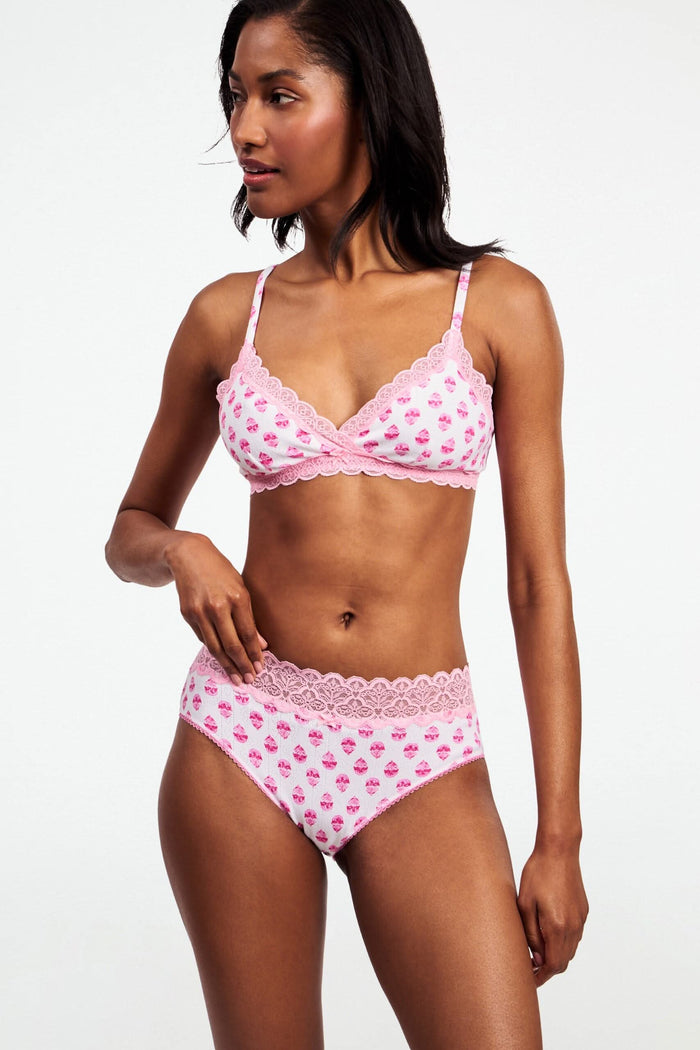 A woman poses sideways in a matching light pink Roller Rabbit Ditsy Daisy Pointelle Cornelia Hipster bra and high-waisted panties with small floral patterns and lace trim, against a plain white background.