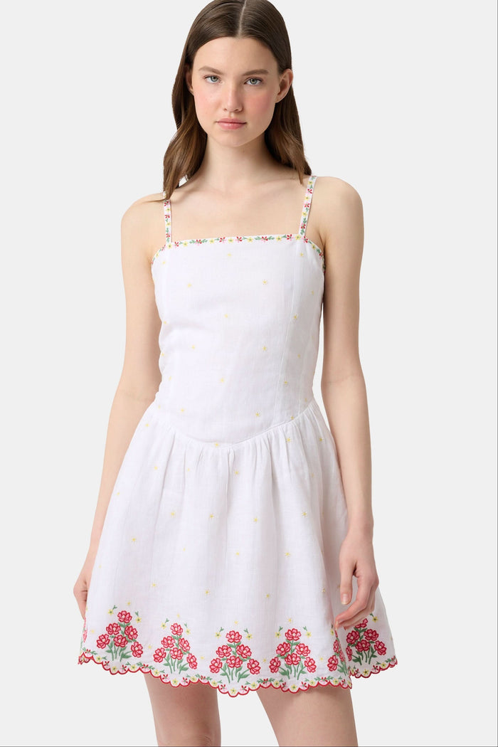 A woman wearing the Roller Rabbit Ditsy Floral Embroidery Hannah Dress—white linen with thin straps, red floral embroidery at the hem and neckline, and yellow flower details—stands against a plain light background.