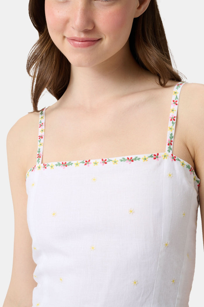 A woman wearing the Roller Rabbit Ditsy Floral Embroidery Hannah Dress—white linen with thin straps, red floral embroidery at the hem and neckline, and yellow flower details—stands against a plain light background.