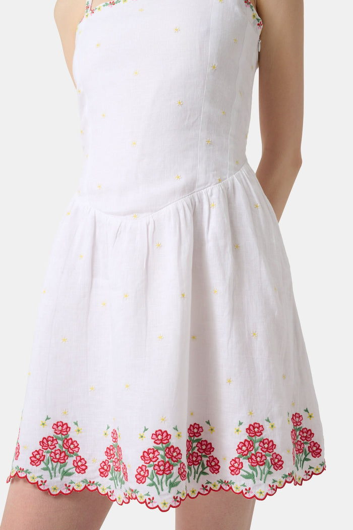 A woman wearing the Roller Rabbit Ditsy Floral Embroidery Hannah Dress—white linen with thin straps, red floral embroidery at the hem and neckline, and yellow flower details—stands against a plain light background.