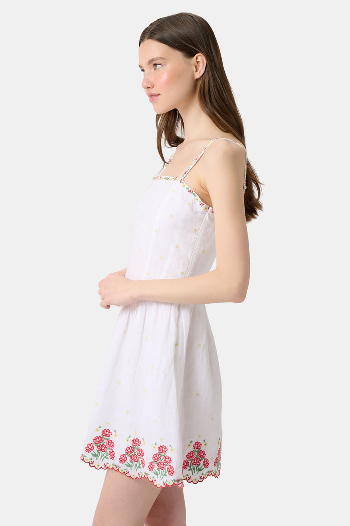 A woman wearing the Roller Rabbit Ditsy Floral Embroidery Hannah Dress—white linen with thin straps, red floral embroidery at the hem and neckline, and yellow flower details—stands against a plain light background.