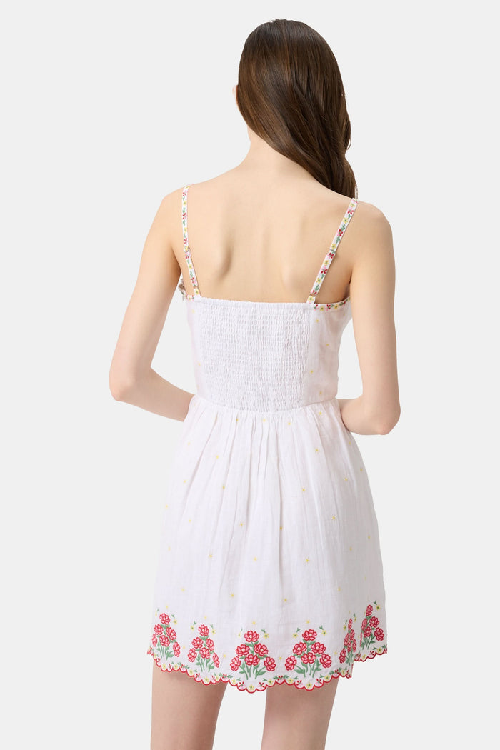 A woman wearing the Roller Rabbit Ditsy Floral Embroidery Hannah Dress—white linen with thin straps, red floral embroidery at the hem and neckline, and yellow flower details—stands against a plain light background.
