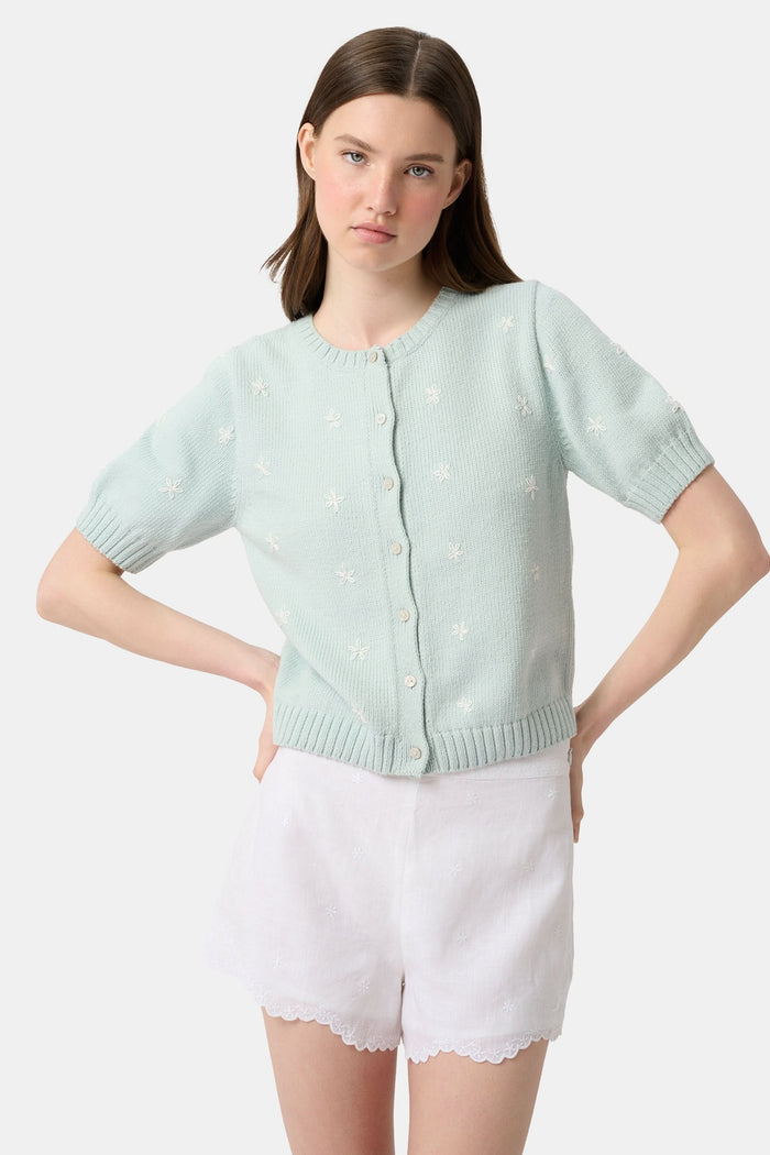 A person wears the Roller Rabbit Ditsy Floral Embroidery Jordana Shorts—white, high-waisted, with a subtle floral design and scalloped lace trim—paired with a light green buttoned sleeveless knit top. The background is plain white.