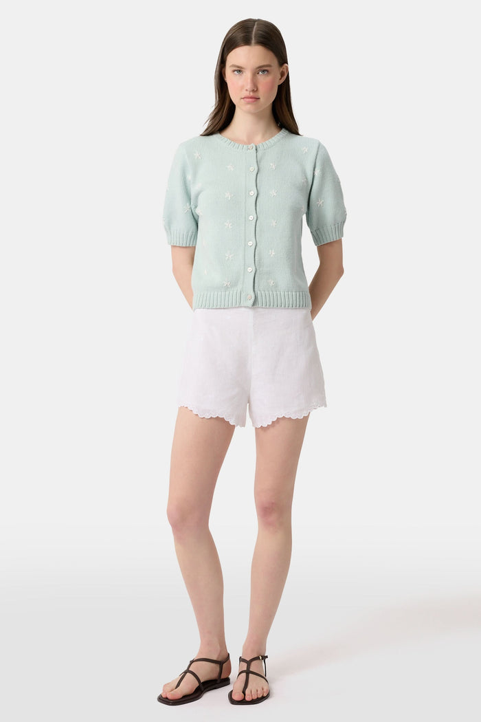 A person wears the Roller Rabbit Ditsy Floral Embroidery Jordana Shorts—white, high-waisted, with a subtle floral design and scalloped lace trim—paired with a light green buttoned sleeveless knit top. The background is plain white.