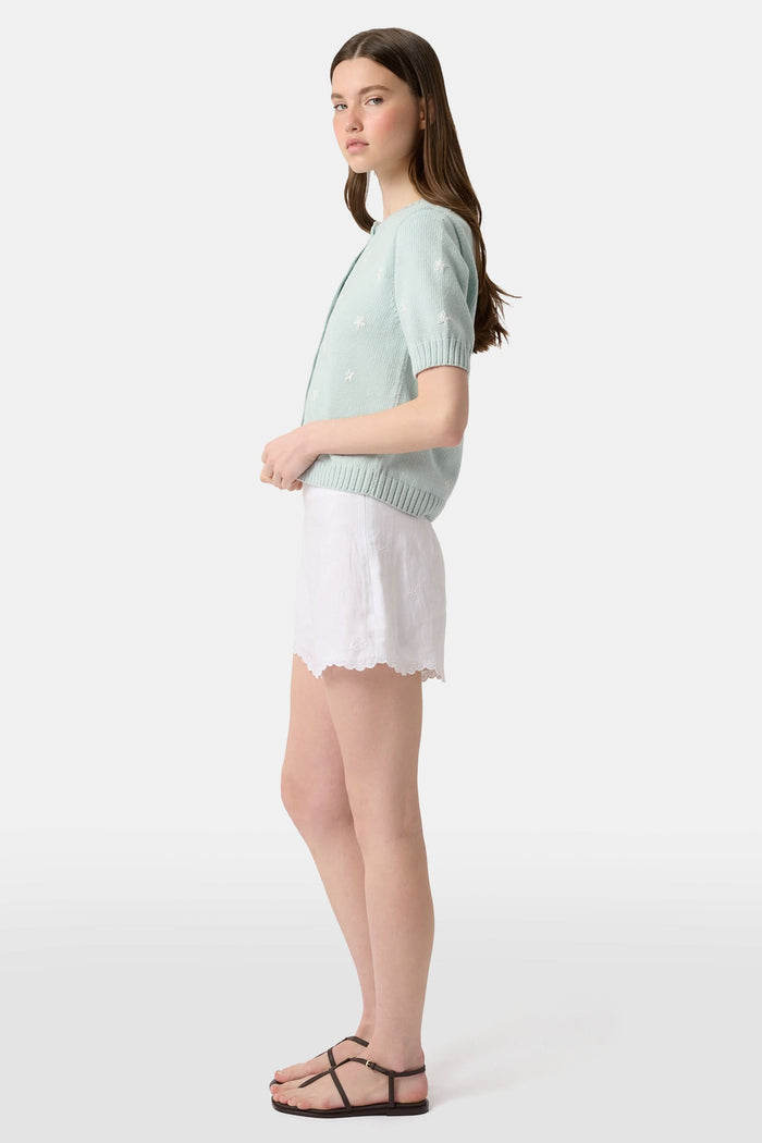 A person wears the Roller Rabbit Ditsy Floral Embroidery Jordana Shorts—white, high-waisted, with a subtle floral design and scalloped lace trim—paired with a light green buttoned sleeveless knit top. The background is plain white.