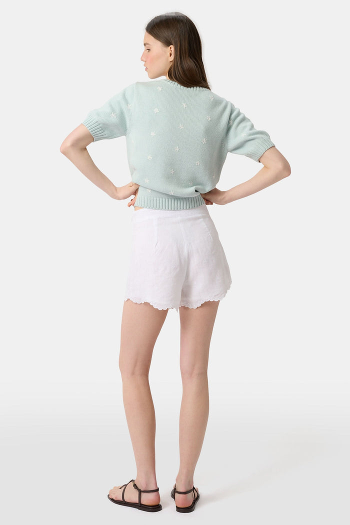 A person wears the Roller Rabbit Ditsy Floral Embroidery Jordana Shorts—white, high-waisted, with a subtle floral design and scalloped lace trim—paired with a light green buttoned sleeveless knit top. The background is plain white.