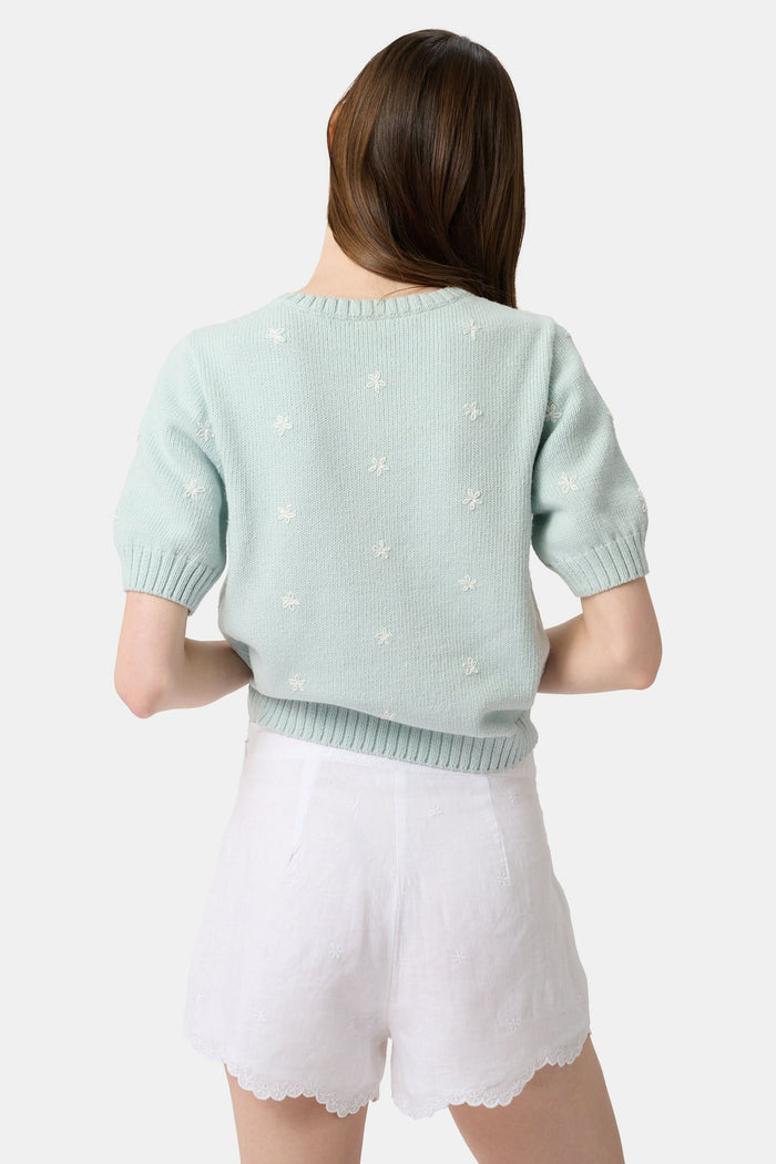 A person wears the Roller Rabbit Ditsy Floral Embroidery Jordana Shorts—white, high-waisted, with a subtle floral design and scalloped lace trim—paired with a light green buttoned sleeveless knit top. The background is plain white.