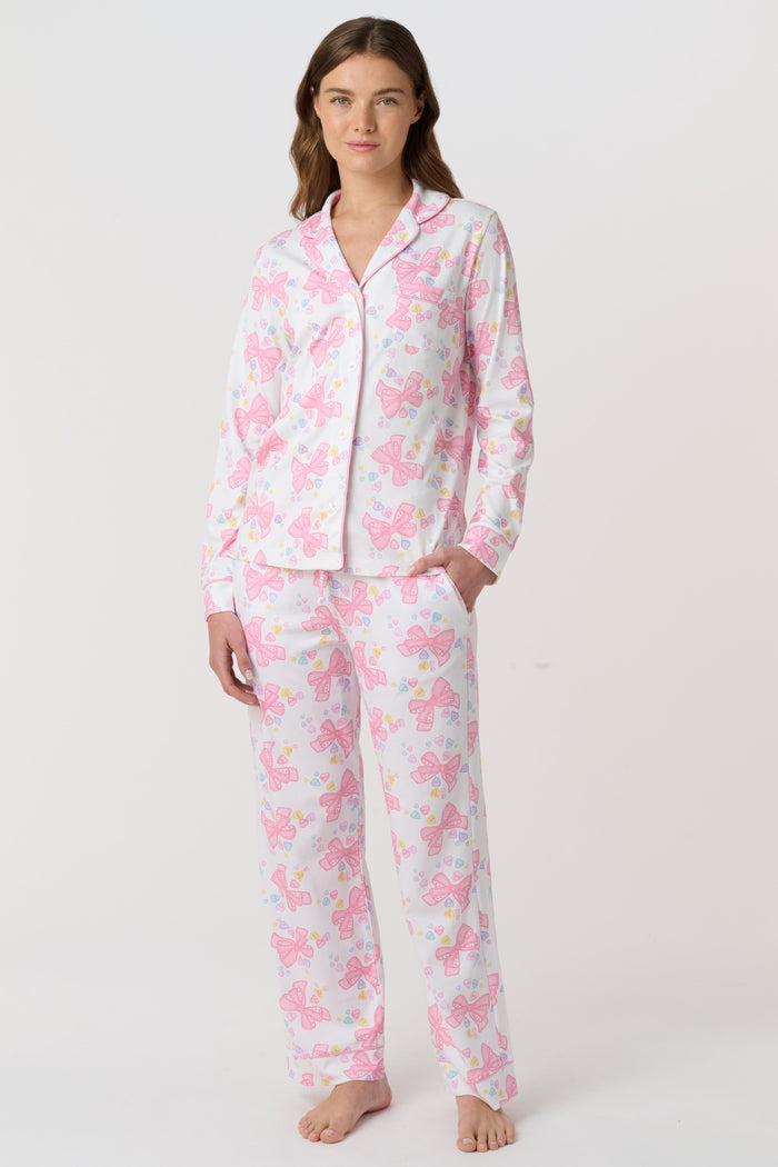A woman stands barefoot, facing the camera in Roller Rabbit Doily Beaux Long Sleeve Polo Pajamas featuring a pink print. The matching set includes a button-up top and long pants; she wears a neutral expression.