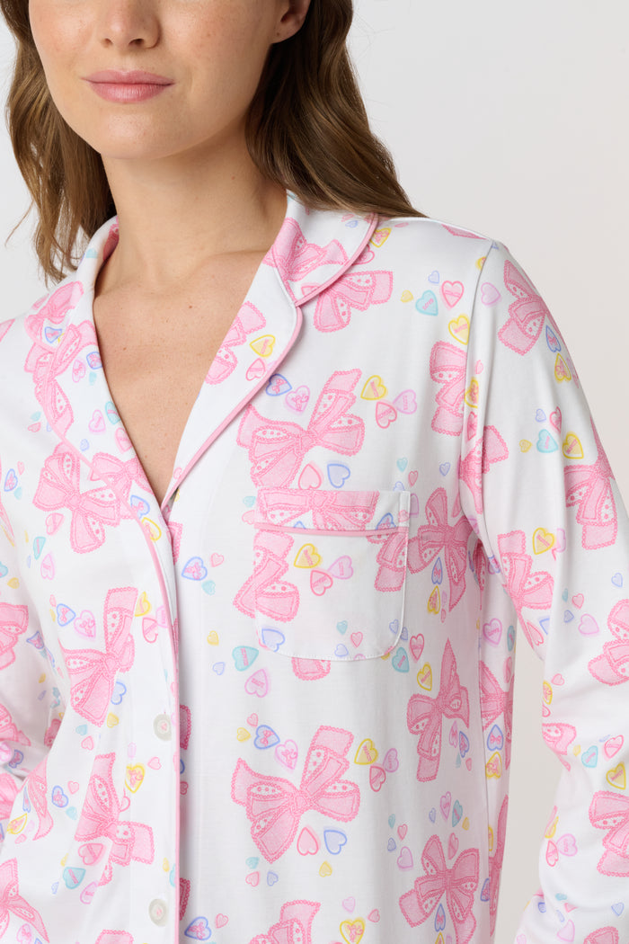 A woman stands barefoot, facing the camera in Roller Rabbit Doily Beaux Long Sleeve Polo Pajamas featuring a pink print. The matching set includes a button-up top and long pants; she wears a neutral expression.