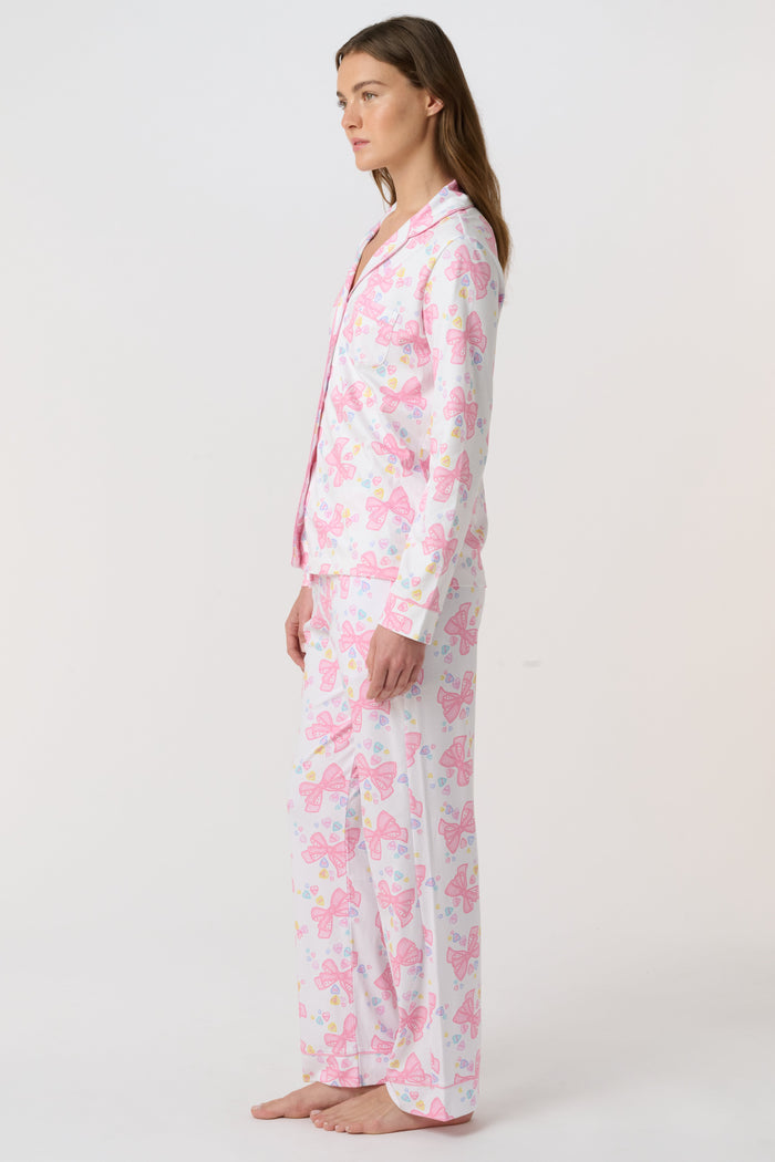 A woman stands barefoot, facing the camera in Roller Rabbit Doily Beaux Long Sleeve Polo Pajamas featuring a pink print. The matching set includes a button-up top and long pants; she wears a neutral expression.