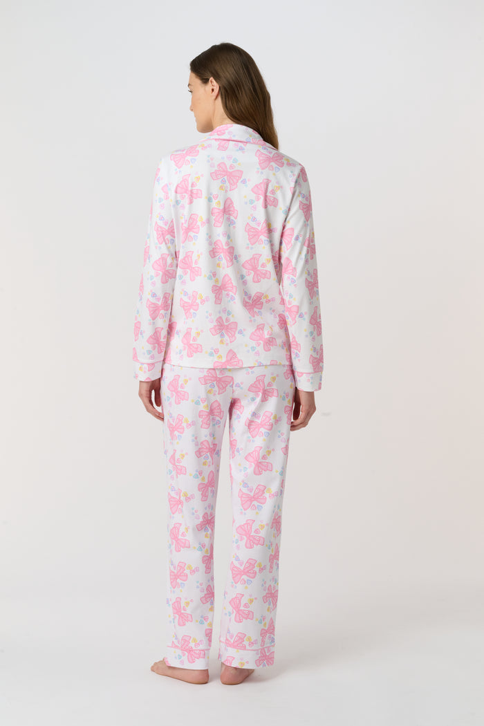 A woman stands barefoot, facing the camera in Roller Rabbit Doily Beaux Long Sleeve Polo Pajamas featuring a pink print. The matching set includes a button-up top and long pants; she wears a neutral expression.