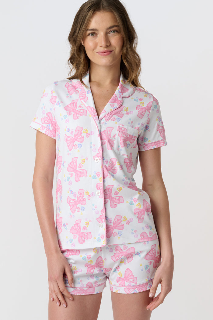 A woman stands smiling against a plain light background, wearing Roller Rabbit’s Doily Beaux Polo Pajamas—a short-sleeved, collared white set with pink bow patterns and small colorful shapes.