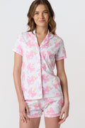 A woman stands smiling against a plain light background, wearing Roller Rabbit’s Doily Beaux Polo Pajamas—a short-sleeved, collared white set with pink bow patterns and small colorful shapes.