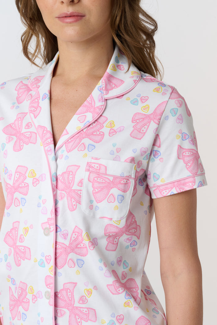 A woman stands smiling against a plain light background, wearing Roller Rabbit’s Doily Beaux Polo Pajamas—a short-sleeved, collared white set with pink bow patterns and small colorful shapes.