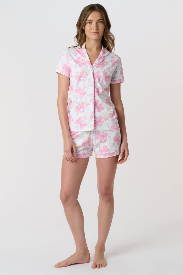 A woman stands smiling against a plain light background, wearing Roller Rabbit’s Doily Beaux Polo Pajamas—a short-sleeved, collared white set with pink bow patterns and small colorful shapes.