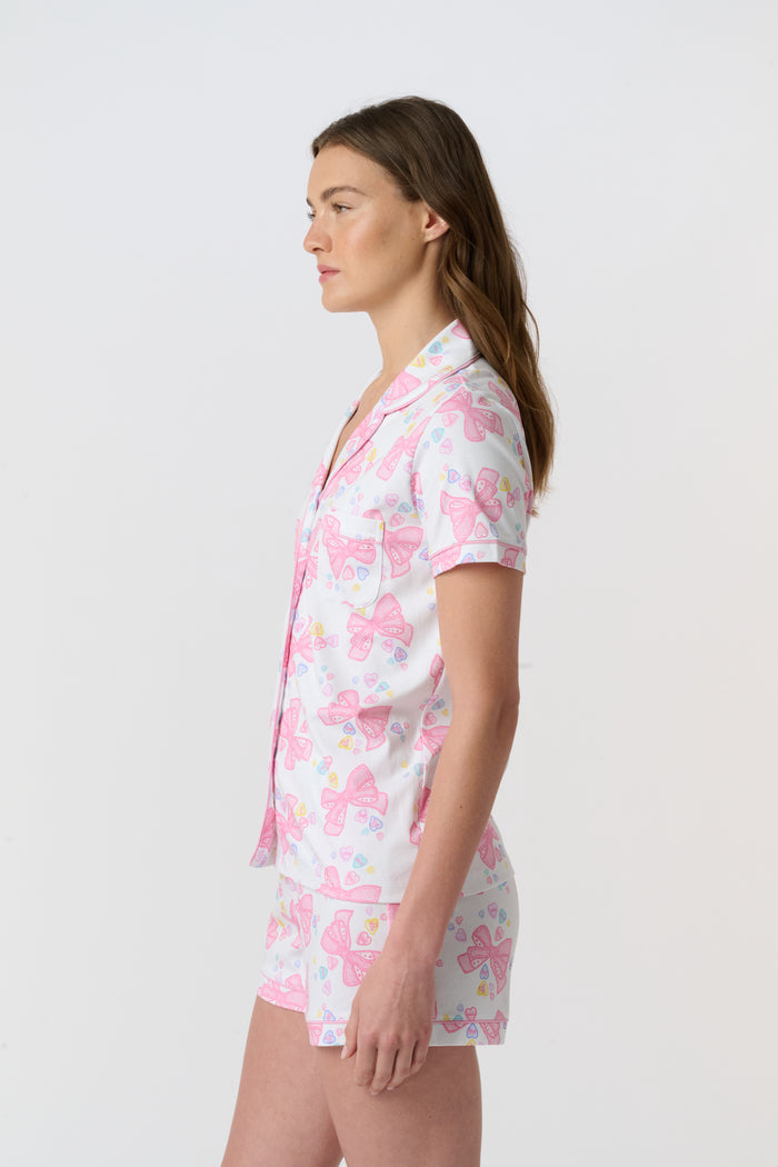 A woman stands smiling against a plain light background, wearing Roller Rabbit’s Doily Beaux Polo Pajamas—a short-sleeved, collared white set with pink bow patterns and small colorful shapes.
