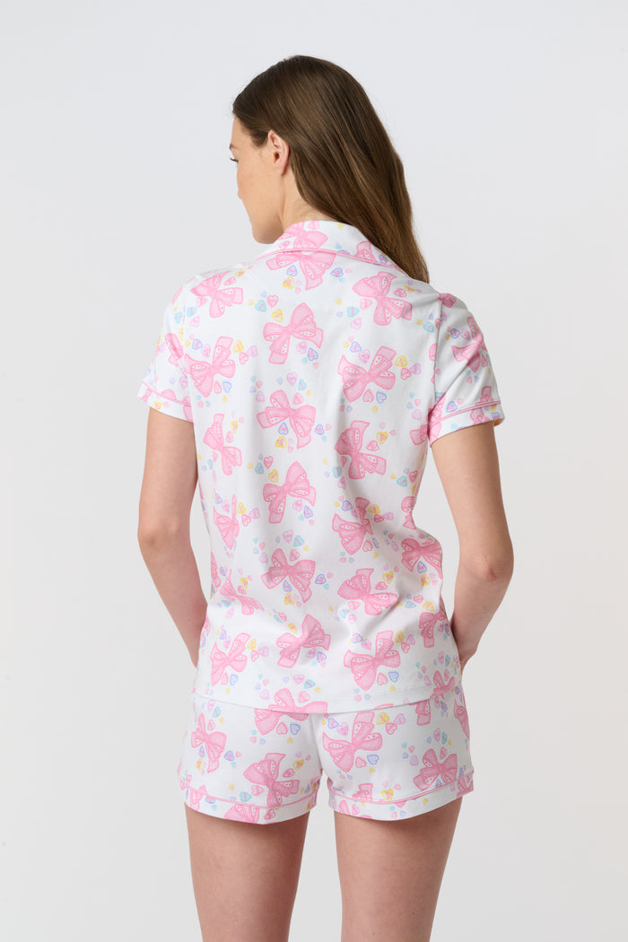 A woman stands smiling against a plain light background, wearing Roller Rabbit’s Doily Beaux Polo Pajamas—a short-sleeved, collared white set with pink bow patterns and small colorful shapes.