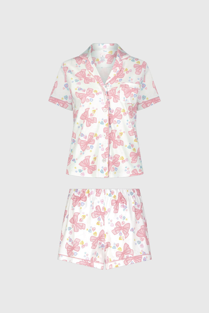 A woman stands smiling against a plain light background, wearing Roller Rabbit’s Doily Beaux Polo Pajamas—a short-sleeved, collared white set with pink bow patterns and small colorful shapes.