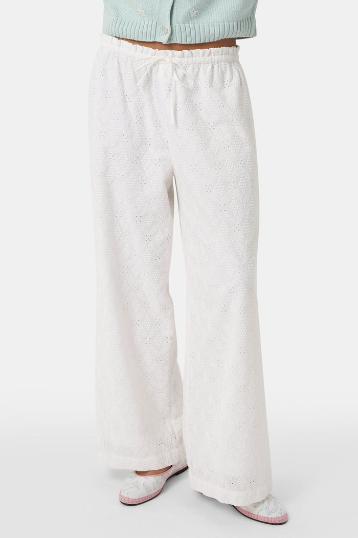 A person wearing Roller Rabbit's Eyelet Lattice Lynette Pant—loose-fitting white cotton with a drawstring waist—paired with a light blue knit cardigan and pink patterned slippers, standing against a white background.