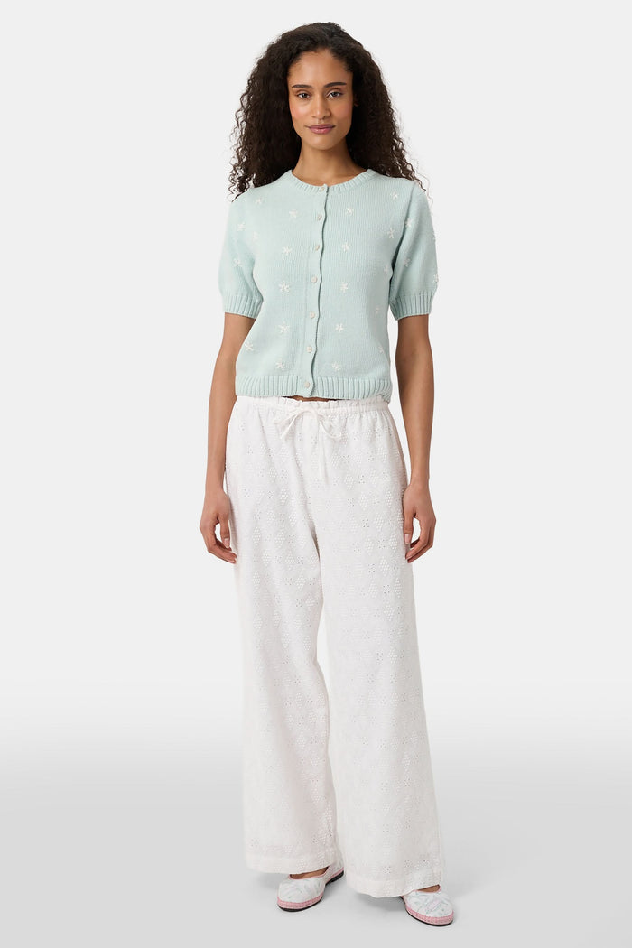 A person wearing Roller Rabbit's Eyelet Lattice Lynette Pant—loose-fitting white cotton with a drawstring waist—paired with a light blue knit cardigan and pink patterned slippers, standing against a white background.
