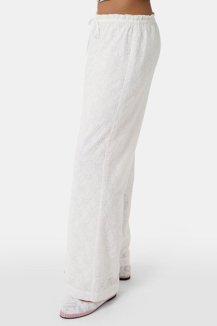 A person wearing Roller Rabbit's Eyelet Lattice Lynette Pant—loose-fitting white cotton with a drawstring waist—paired with a light blue knit cardigan and pink patterned slippers, standing against a white background.