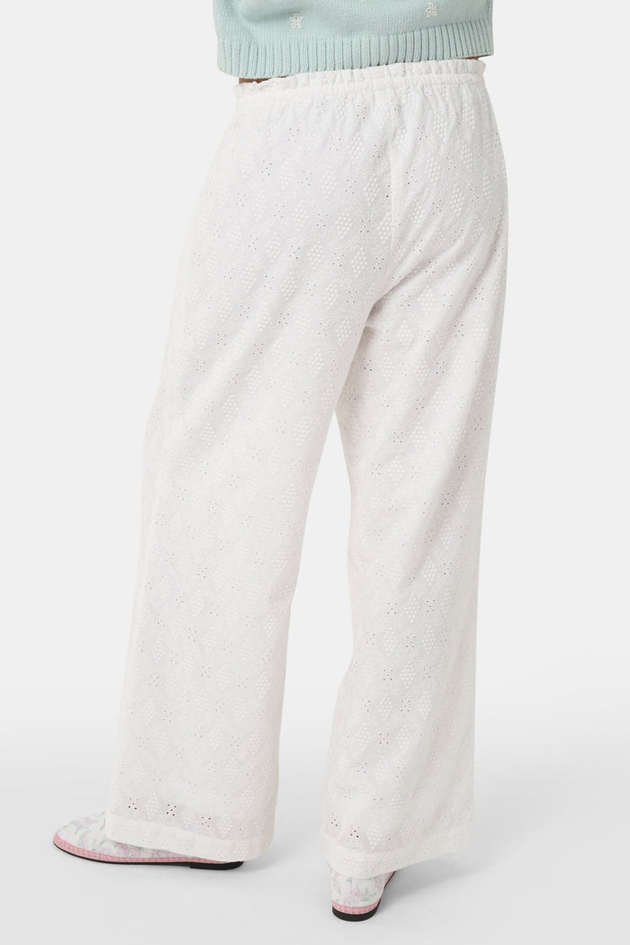 A person wearing Roller Rabbit's Eyelet Lattice Lynette Pant—loose-fitting white cotton with a drawstring waist—paired with a light blue knit cardigan and pink patterned slippers, standing against a white background.