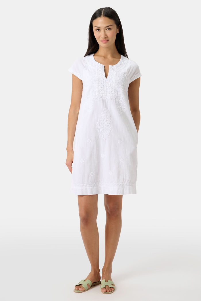 A woman stands against a plain light background, wearing the Roller Rabbit Faith Dress—a short-sleeve, knee-length white embroidered dress with a keyhole neckline—paired with green sandals.