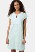 A woman with long curly hair wears the Roller Rabbit Faith Dress, a seaglass-green short-sleeved dress with hand embroidery and a notched neckline, standing against a plain light background.