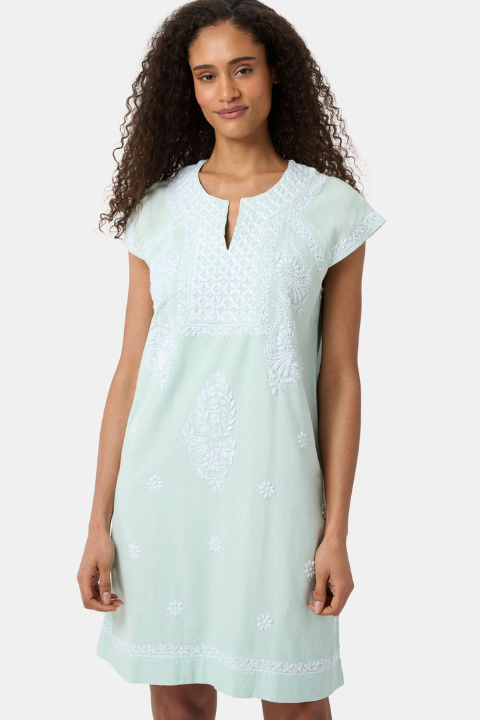 A woman with long curly hair wears the Roller Rabbit Faith Dress, a seaglass-green short-sleeved dress with hand embroidery and a notched neckline, standing against a plain light background.
