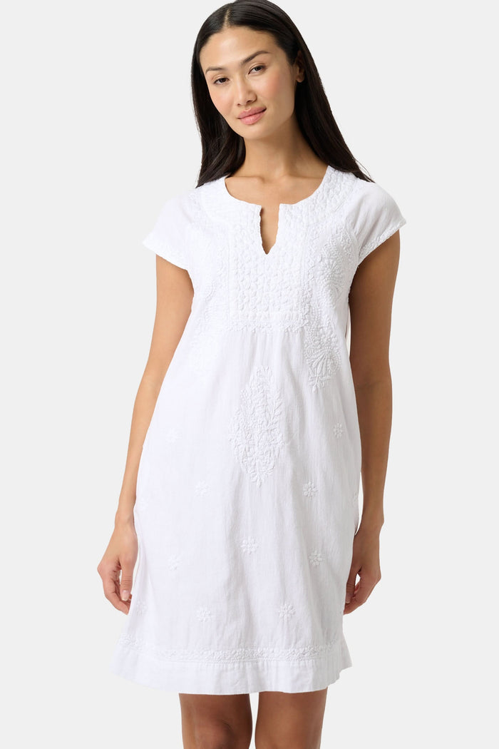 A woman stands against a plain light background, wearing the Roller Rabbit Faith Dress—a short-sleeve, knee-length white embroidered dress with a keyhole neckline—paired with green sandals.