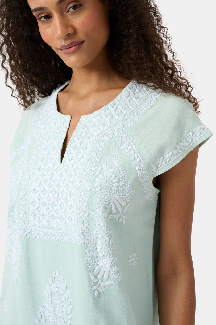 A woman with long curly hair wears the Roller Rabbit Faith Dress, a seaglass-green short-sleeved dress with hand embroidery and a notched neckline, standing against a plain light background.