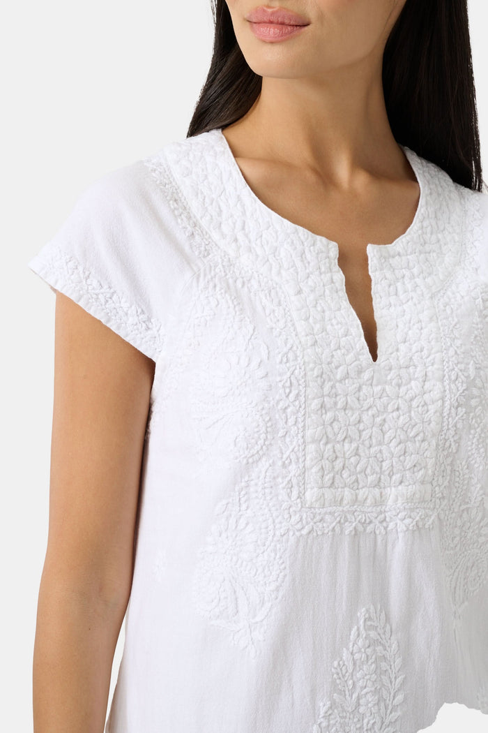 A woman stands against a plain light background, wearing the Roller Rabbit Faith Dress—a short-sleeve, knee-length white embroidered dress with a keyhole neckline—paired with green sandals.